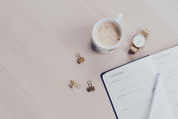 A foamy latte, binder clips, a watch, and an open planner all sit on a wooden table.