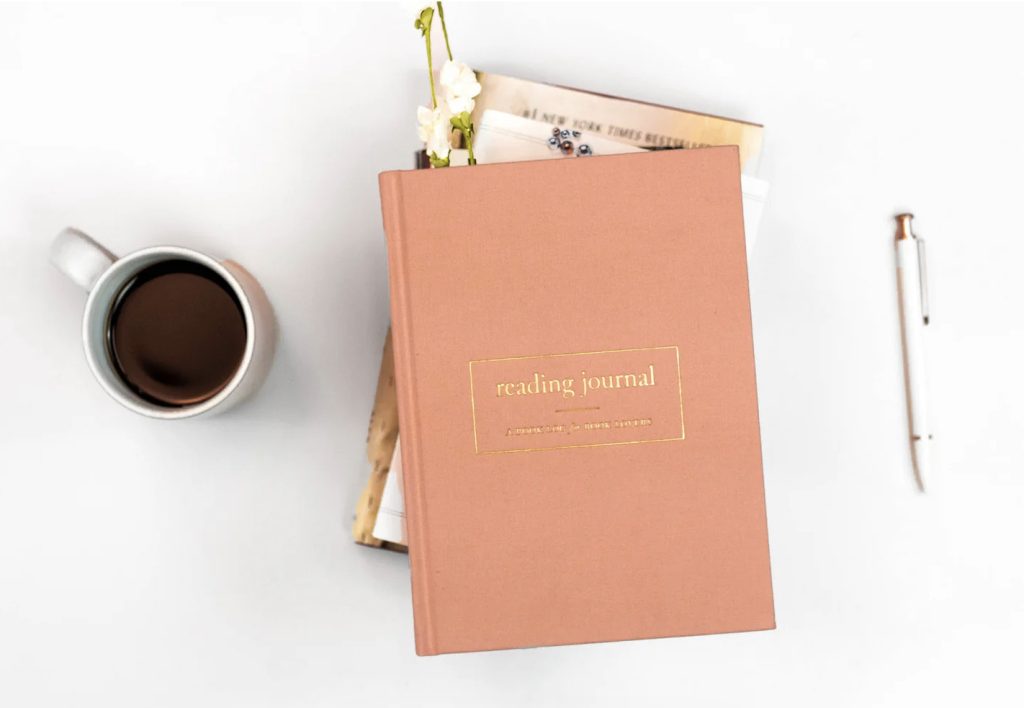 A salmon pink reading journal sits with gold lettering atop a stack of books beside a cup of coffee and a white pen.