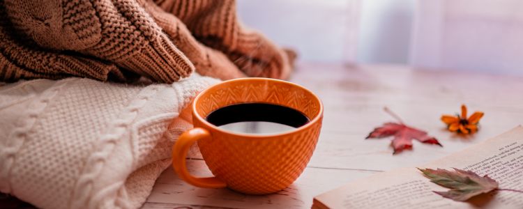 knit sweaters stacked to the left, orange mug filled with coffee in the center, and book pages and fall leaves to the right