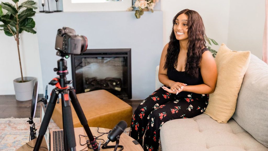 A woman filming a video while sitting on a tan couch. In front of her is a camera on a tripod, a microphone, a laptop, and a ring light.