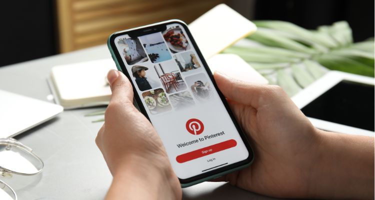 A person at a desk holds up a phone that has a "Welcome to Pinterest" screen pulled up.