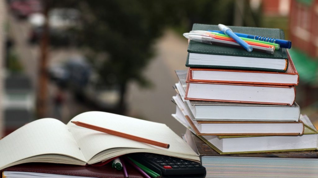 There is a stack of books with pens on top. Next to it lies an open notebook with a pencil and calculator underneath.