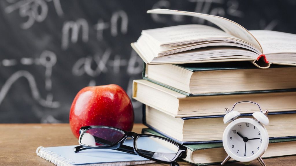 There is a table with a stack of books, an apple, notepad, glasses, and a clock. In the background is a chalkboard with equations on it.