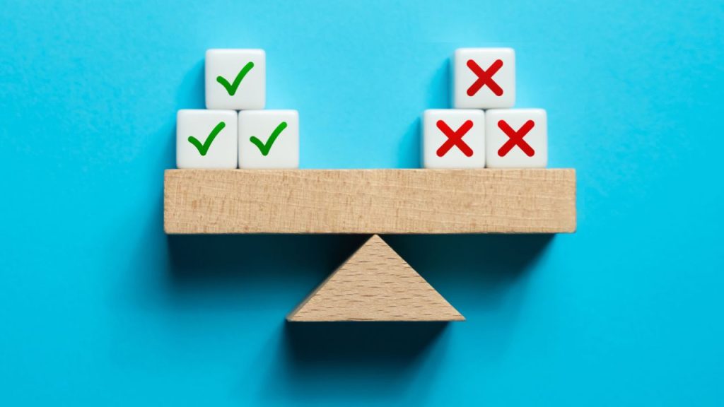 A wooden block scale with three white cubes with green checks mark on them on the left side and three white cubes with red X marks on them on the right.