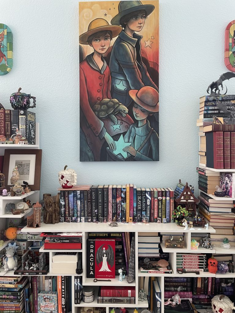 Image of Kiersten White's bookshelf filled with books and knickknacks with a large painting of two women and one young girl holding stars and a turtle beside them.
