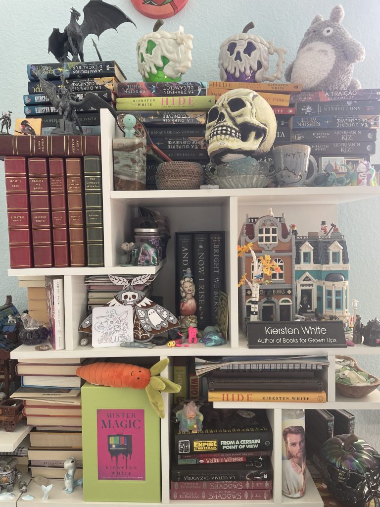 A closer look at Kiersten White's bookshelf with books and knickknacks, mainly Halloween-themed. On the top right of the bookshelf lies five of her own books with custom leatherbound covers.
