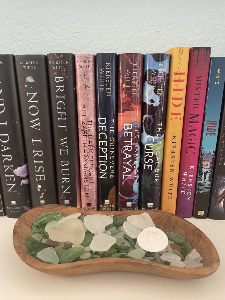 Image of a collection of some books by Kiersten White on her shelf with a wooden bowl of beach glass in front of it.