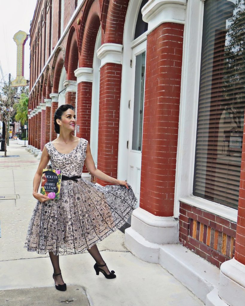 Carmen wearing a vintage dress holding a book standing in front of a red brick building