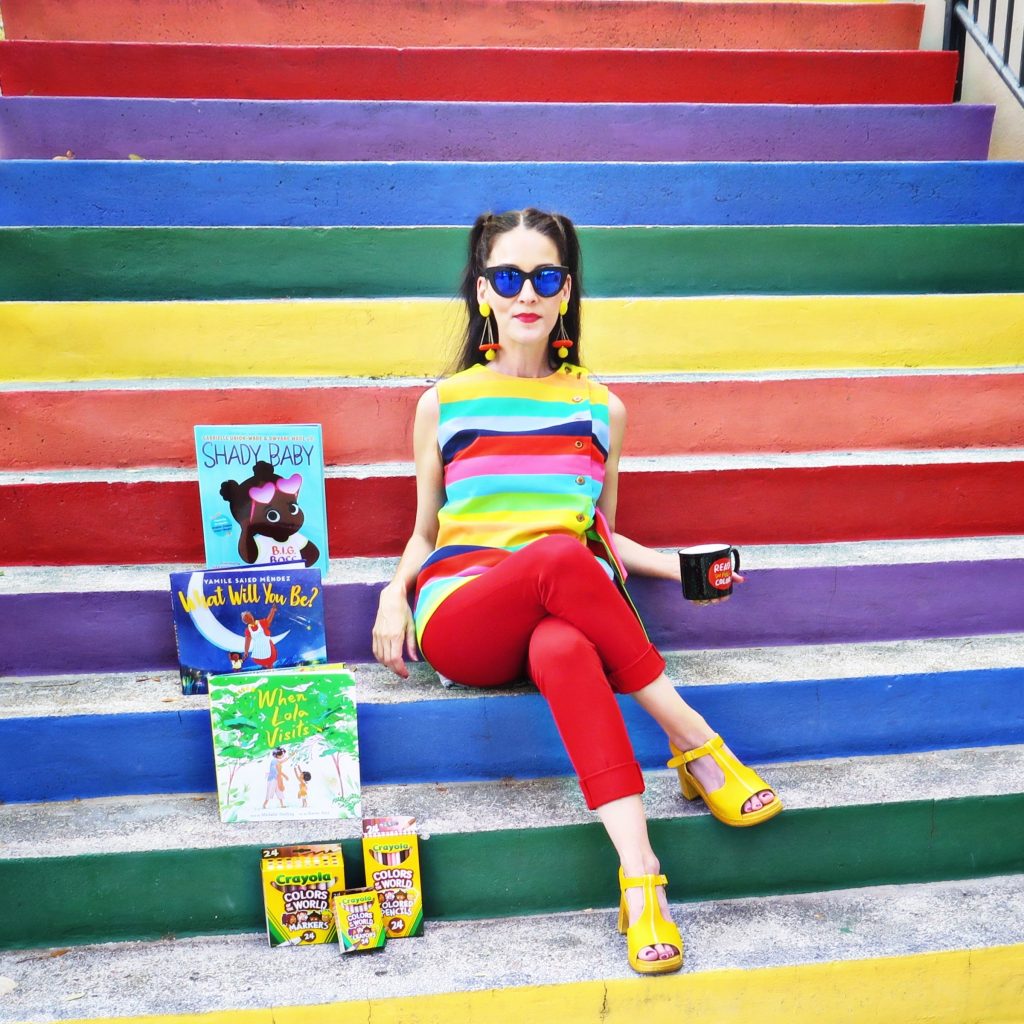 Carmen sitting on rainbow stairs with children's books and crayons while holding a mug
