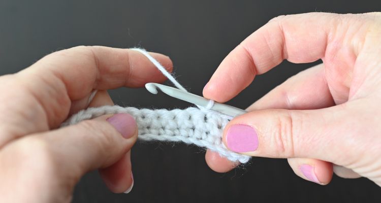 Hands with pink fingernails hold a silver crochet hook and a partial project of white yarn.