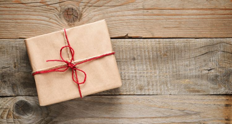 A rectangular present with tan wrapping paper sits atop wooden planks. The present has red string in a bow on it.