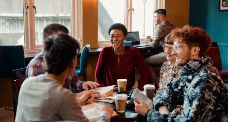 A group of people are sitting in a circle in a cafe looking at books with coffee cups on the table.
