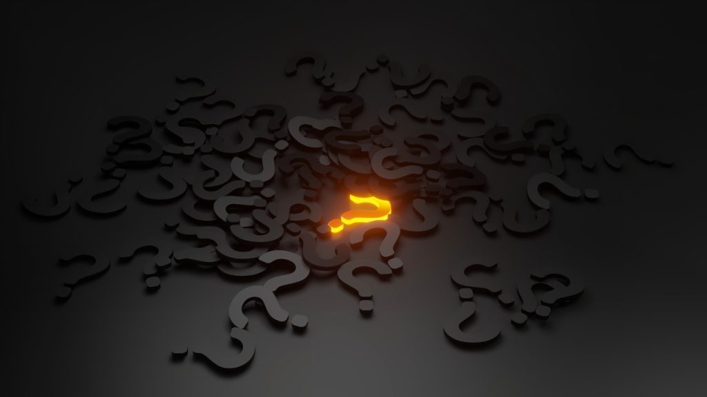 Pile of black question marks on a black table with one bright orange question mark in the middle of the pile.