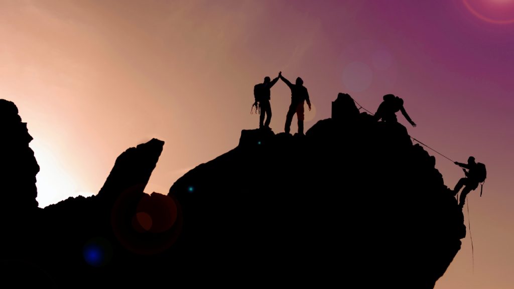 Four people climbing a mountain. One person is climbing via rope and the person above them has their hand extended to help pull them up.