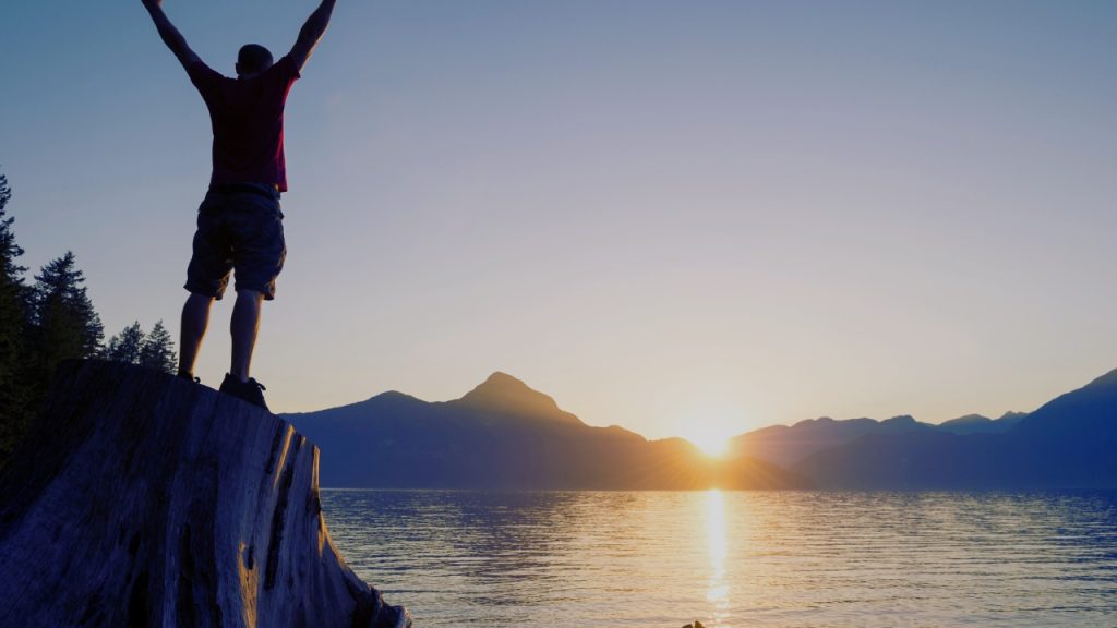A man stands facing a body of water with his arms raised in victory. The sun is setting across the water and behind some mountains, and the reflection is shining across the water.