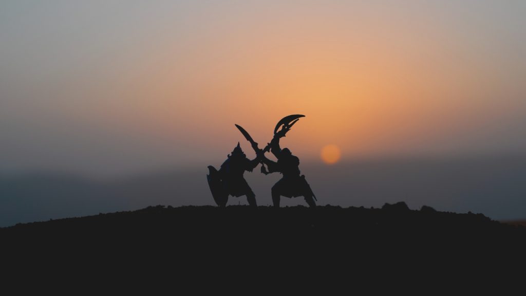 Two men in elaborate armor are silhouetted against a setting sun are fighting with intricately shaped weapons.
