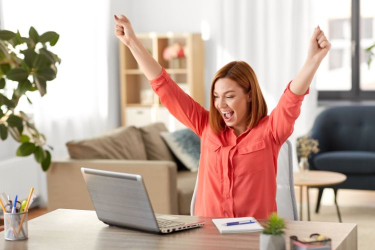 Picture shows woman sitting in front of laptop smiling and with her arms up to celebrate.