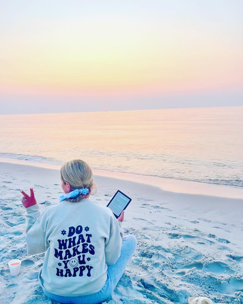 Michelle sitting on the beach infront of the ocean with a kindle in her hand. She has on a shirt that states "Do What Makes You Happy."
