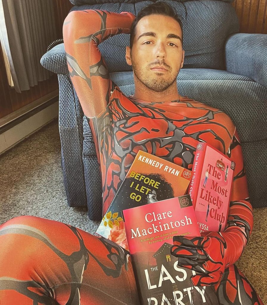 Image of Bookstagrammer Frank DiLuzio in a red and grey superhero costume as he sits on the ground, leaning on his blue, corduroy recliner. He is tilting his head back onto one hand and holding three books with the other hand.
