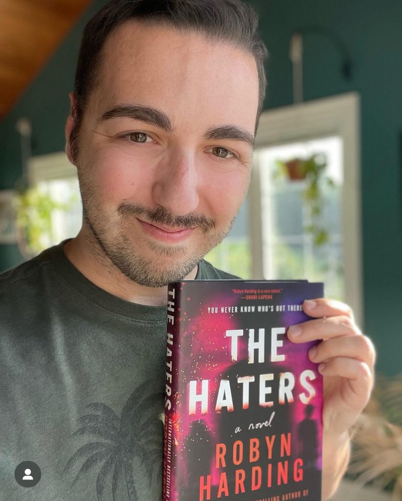 Bookstagrammer Frank DiLuzio holds closely to him the book The Haters by Robyn Harding and he smiles to the camera.