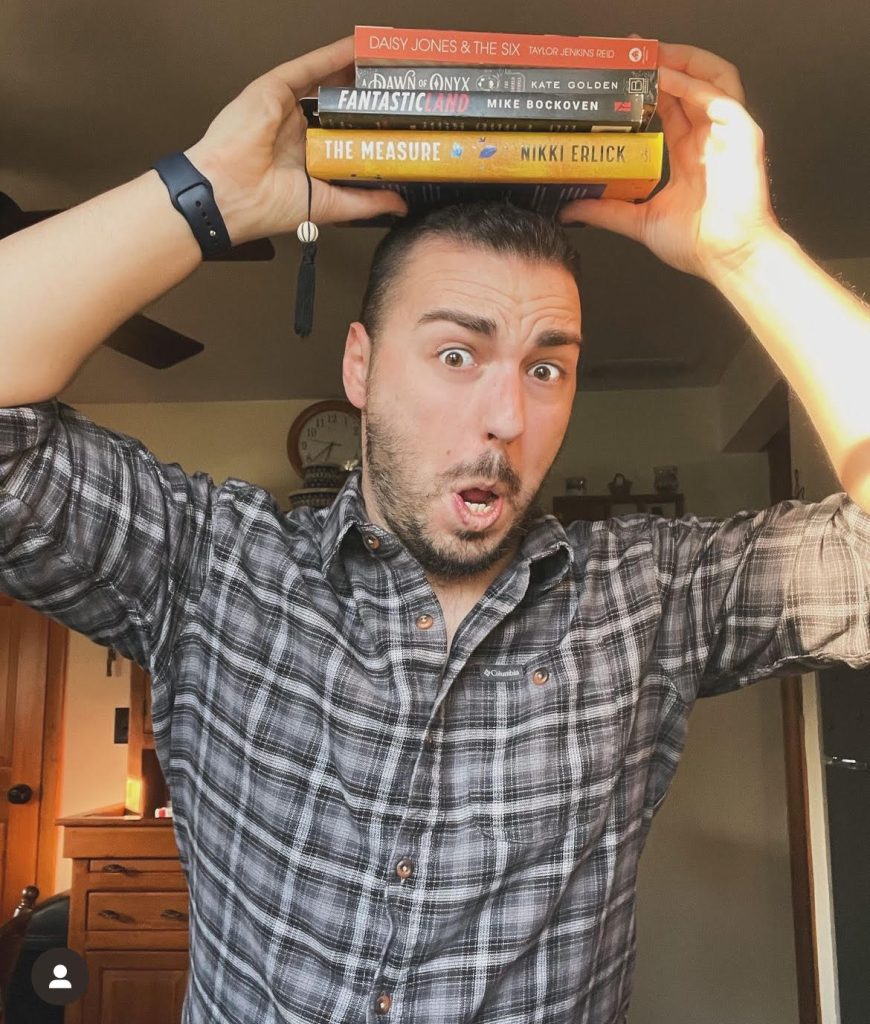 Image of Bookstagrammer Frank DiLuzio scrutching his nose and moth together with his eyebrows raised as he holds a stack of books over his head.