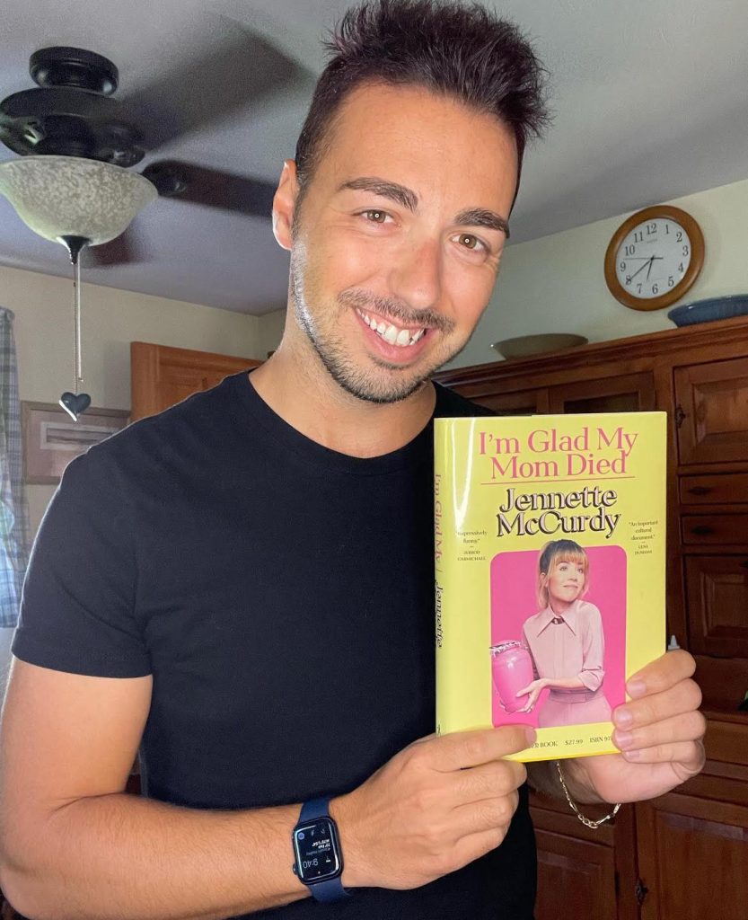 Bookstagrammer Frank DiLuzio holding the book I'm Glad My Mom Died by Jennette McCurdy as he smiles.