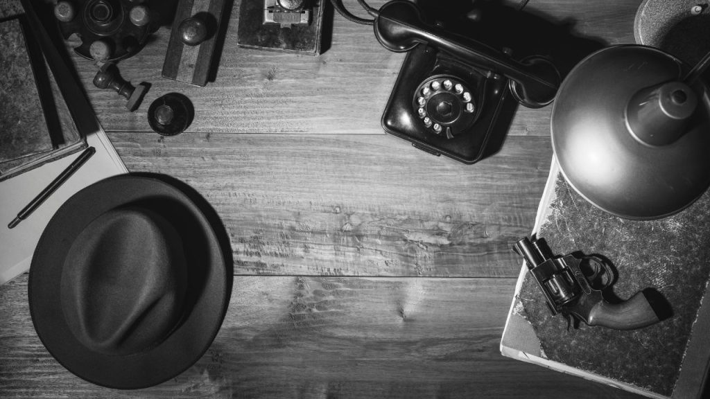 An image of a black and white noir esq style of a table filled with many objects, like a detective's office. A fedora, a gun, an old telephone, and a lamp is on the wooden table.