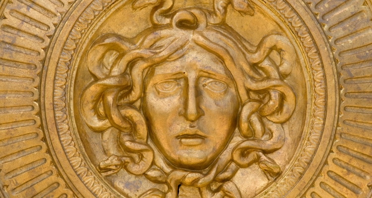 A close-up photo of a gold metal carving of an upset woman's face with a strong nose and snakes instead of hair.