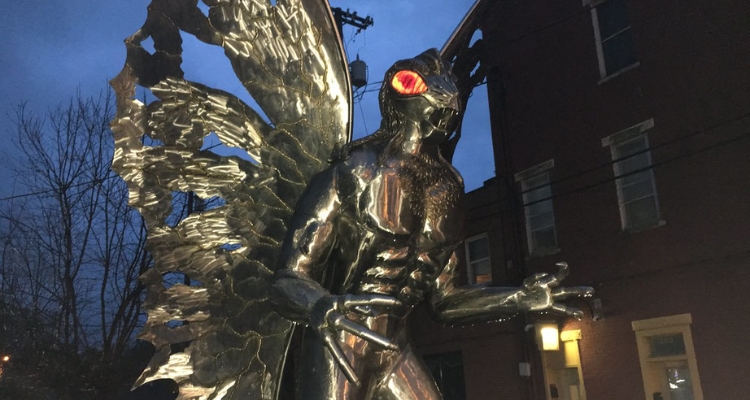 A close-up photo of a large metal statue of a creature with glowing red eyes, moth wings, long claws, and abs.
