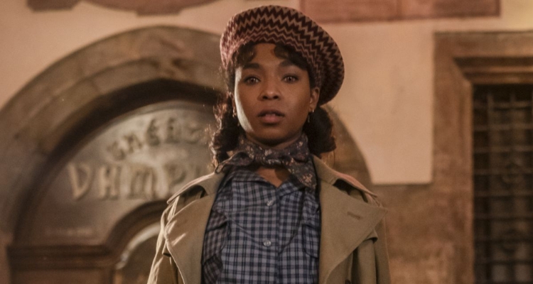 A television still of a young black woman with light brown eyes and curled dark hair wearing a checkered blouse, tan coat, floral neck scarf, and patterned hat.