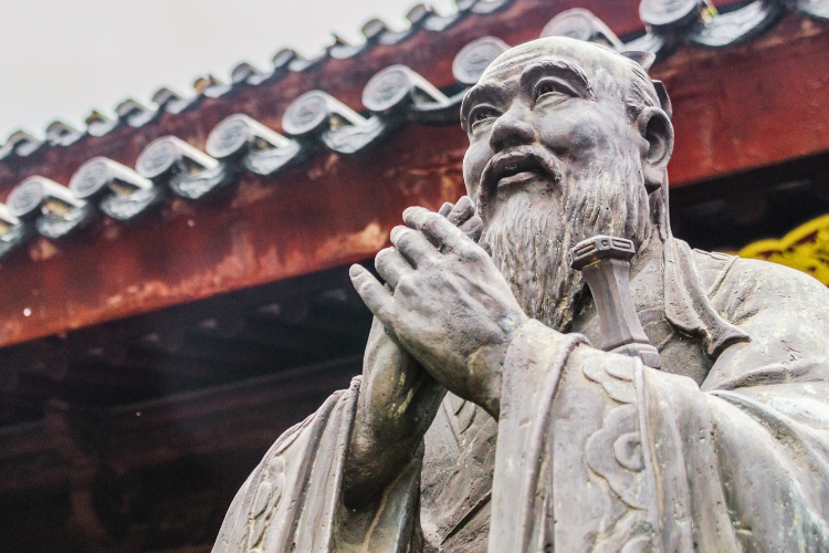 A statue of Confucius with his hands clasped loosely in front of his chest as he looks out into the distance with a contemplative gaze.