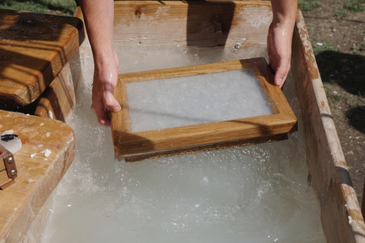 Two hands hold a wooden frame in a container of cloudy water. They are lifting the frame out of the water after having collected some of the paper sediment from the water. Depicts early paper making techniques.