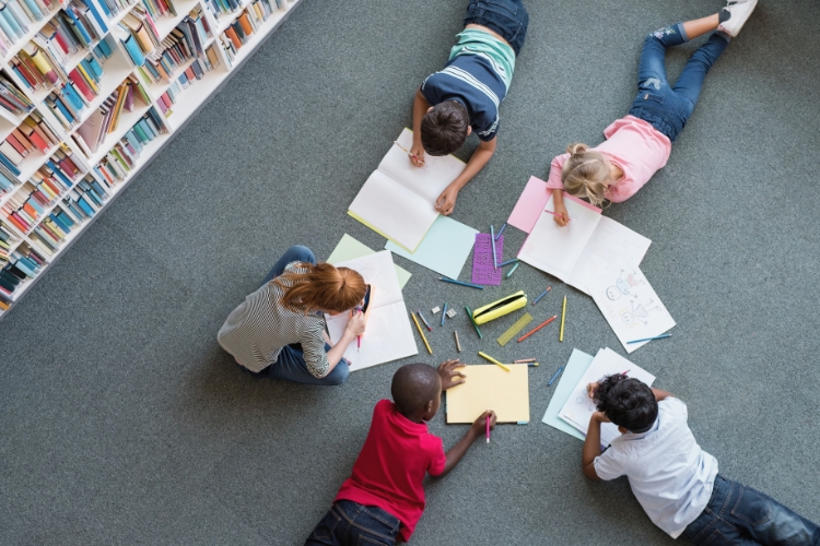 A group of children sit and lay on the grey carpeted floor of a library with various writing utensils and notebooks between them.