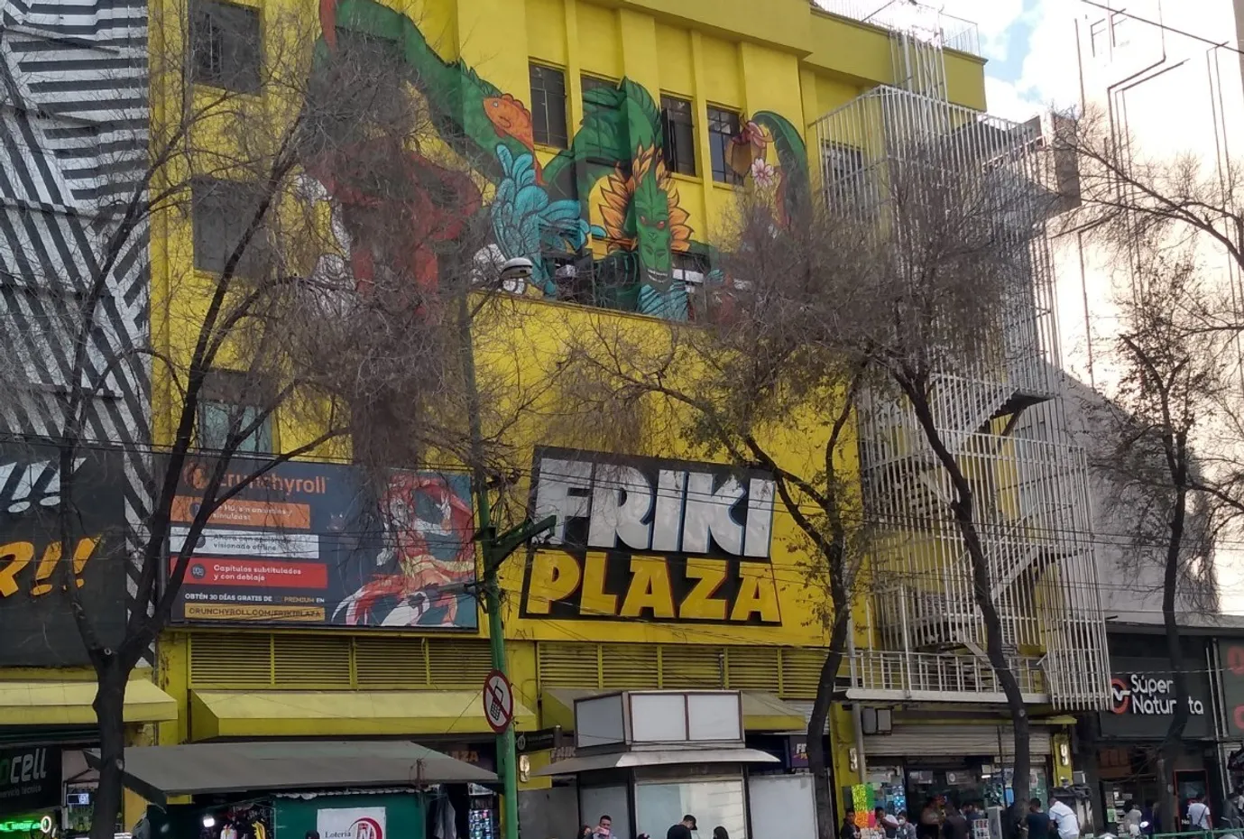 Image of Friki Plaza in Mexico City. A four-story yellow building with the dragon from Dragon Ball painted over a yellow wall. A billboard promoting Crunchyroll is seen next to the building.