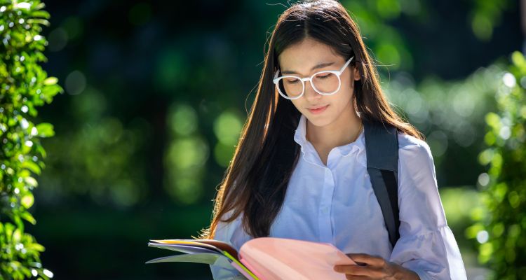 A woman with white glasses reading from a folder of colored paper surrounded by green hedges.