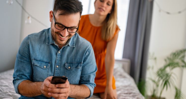 A man laughing on his phone while a woman on the bed behind him tries to look at his phone.
