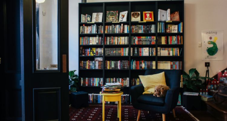 A large black bookshelf in the back of a room, with a blue armchair and a yellow side table in front of it.