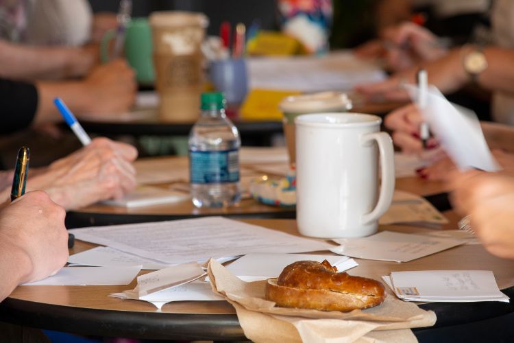 At an up-close table, people are writing on paper. There is a croissant and a few drinks.