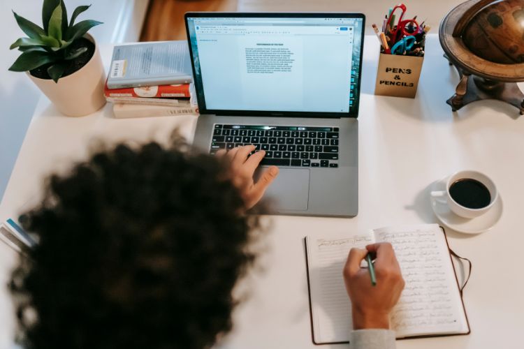 An individual sits writing in a journal at a desk while looking at a laptop screen. There is a small plant, a pile of books, a pencil holder, a globe, and a mug of coffee.