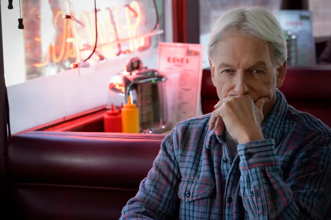 Gibbs sitting at a diner while staring ahead. He is covering his mouth and looking pensively at the camera, wearing a blue flannel.