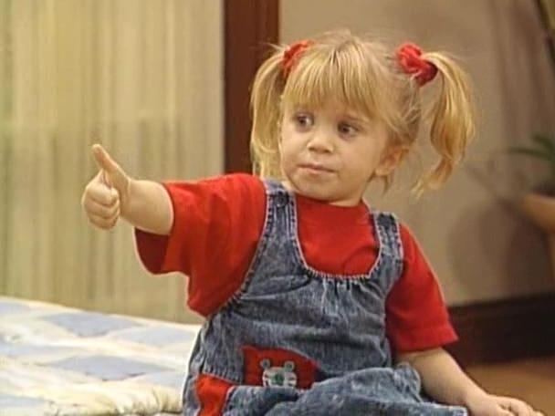 Michelle Tanner giving a thumbs-up. She is wearing a bright red top with denim overalls.