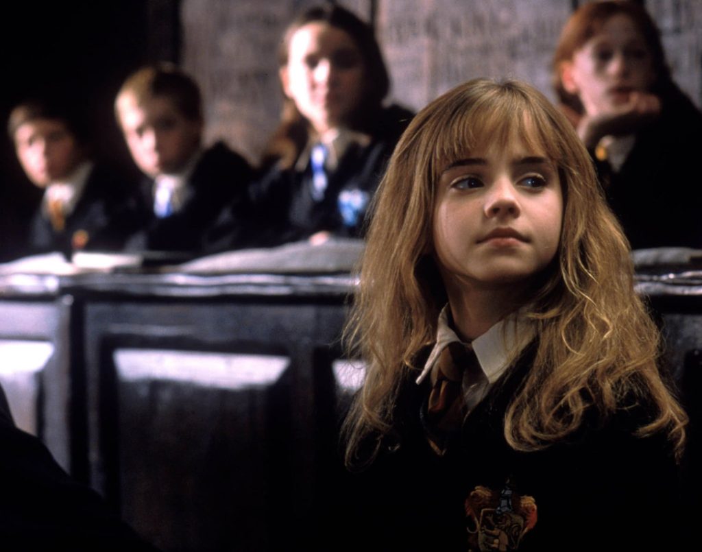 A young girl with dirty blonde hair is wearing a black cloak and tie. She is looking off into the distance and the students behind her are blurred.