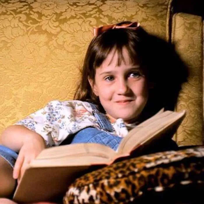 A young girl with brown hair sits on a golden upholstered chair with a book in her hands and a smirk on her face.
