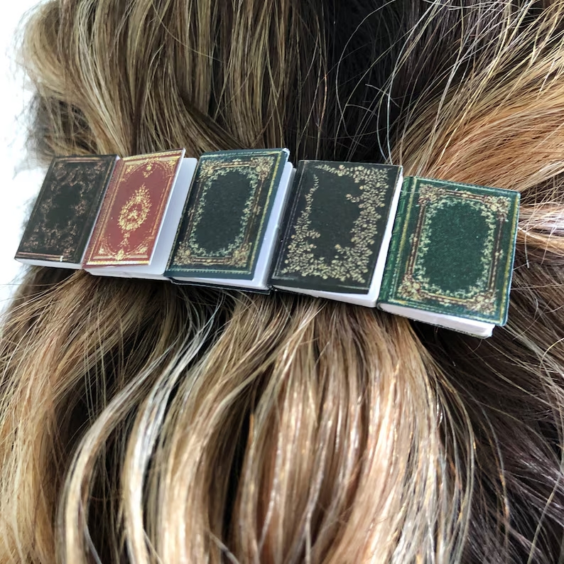 A woman with dark brown hair and blonde highlights has a small barrette holding her hair together. The barrette consists of 5 mini books.