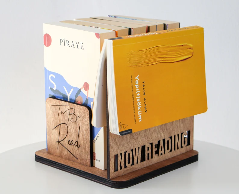 There is a wooden book holder with a mustard yellow book sitting atop the "now reading" engravement. Five paperback books are stacked vertically in the "To Be Read" section.