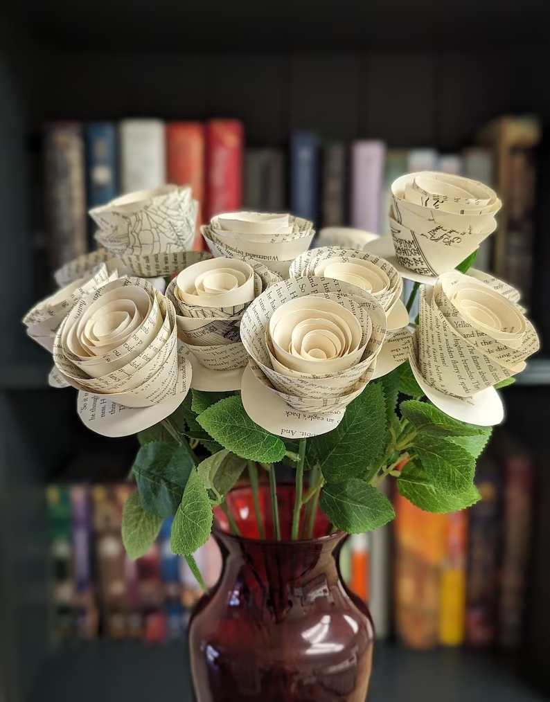 A dozen book page flowers are held in a maroon vase with a blurred out bookshelf in the background.