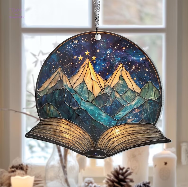 A stained glass ornament with a blue mountain-scape and an unfurling book at the bottom is held in front of a well lit window.