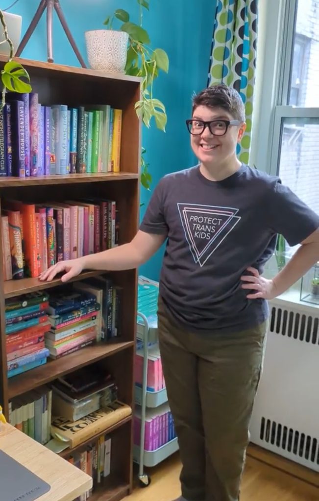 TJ Alexander standing before their book shelf