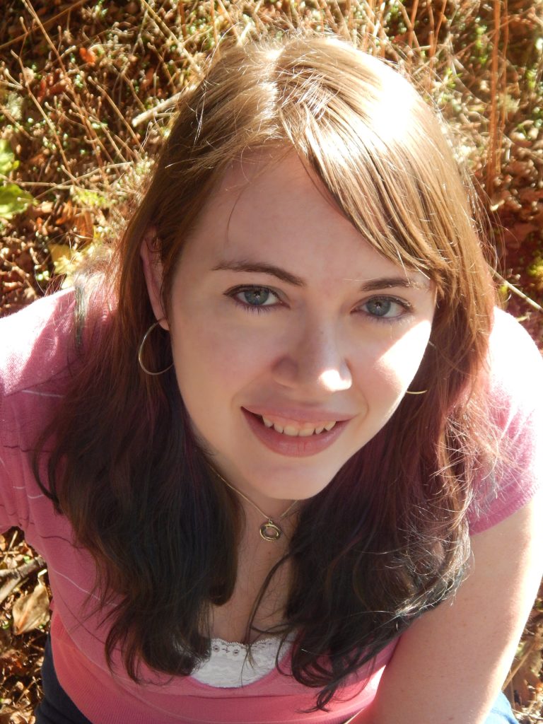 Lyssa in front of a background of what appears to be a ground of grass, with Lyssa wearing a pink and white shirt, styling a pair of hoops and a necklace.