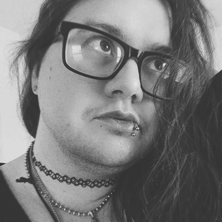 Shane posing, looking off to the side, styling a pair of glasses, choker necklaces, and a lip ring, with hair styled a bit in their face.